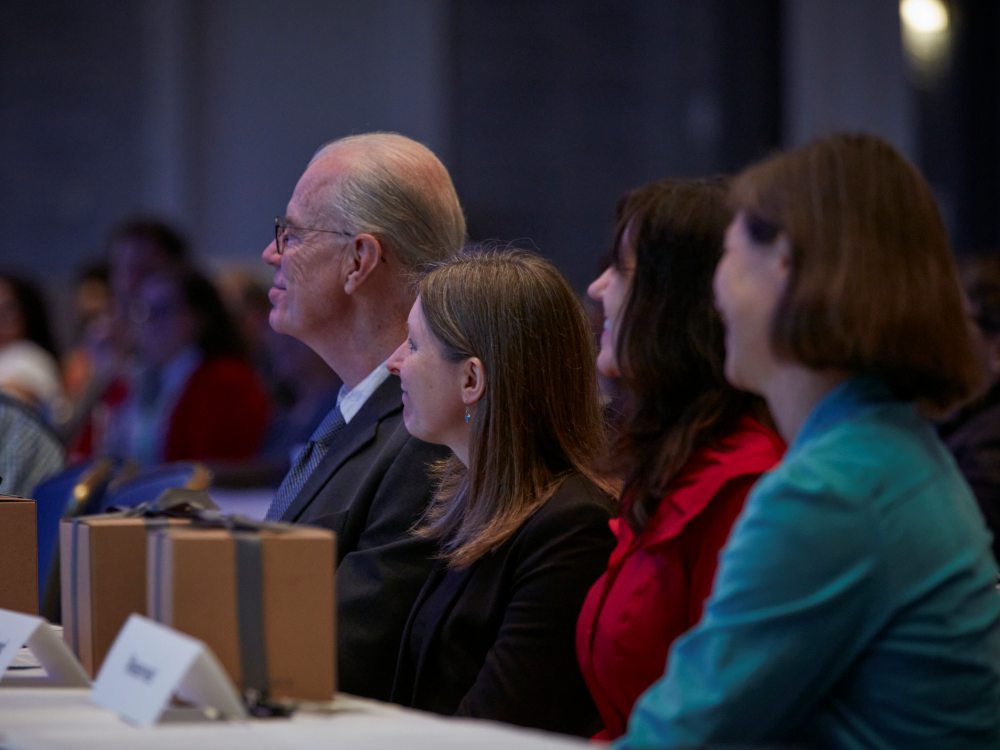 Four people are shown in profile, faces turned away from the camera and seated in a row at a table. On table are three brown gift boxes tied with gray ribbon. Blurred crowd in background. 