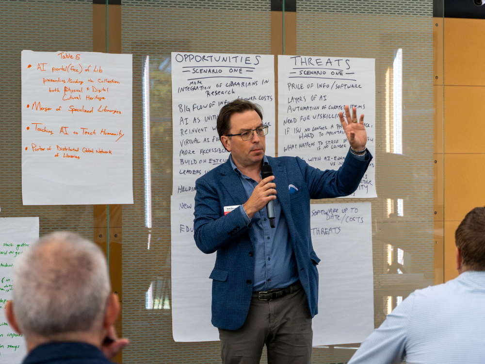 A man speaks into a microphone, standing in front of large sheets of paper containing notes under the headings “Table 5” “Opportunities” and “Threats”