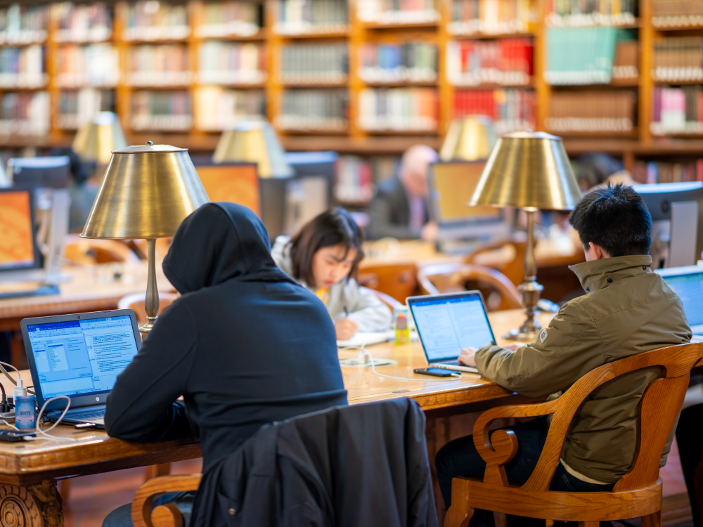 Three young people studying on laptops at a long table with bookshelves in the background.