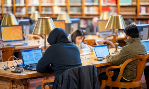 Three young people studying on laptops at a long table with bookshelves in the background.