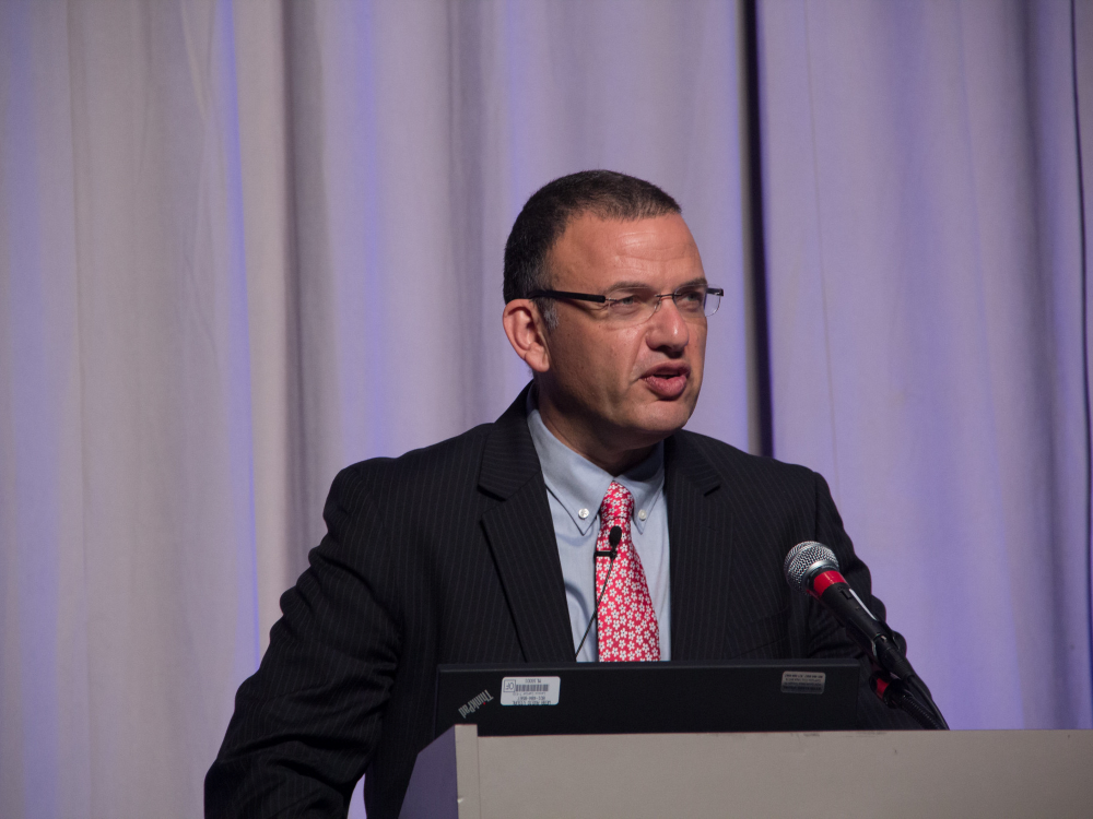 Matti Shem Tov standing at a podium in front of a curtain 