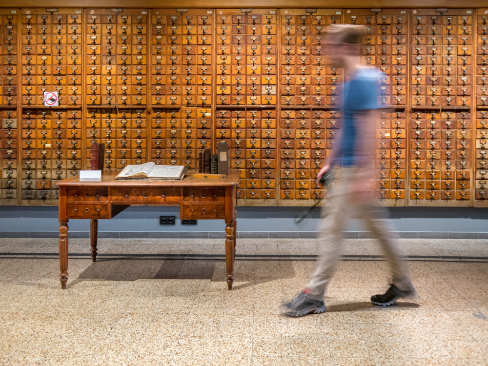 A man, blurred, walks past a massive wooden card catalogue