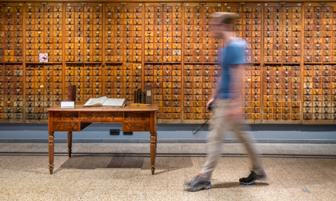 A man, blurred, walks past a massive wooden card catalogue