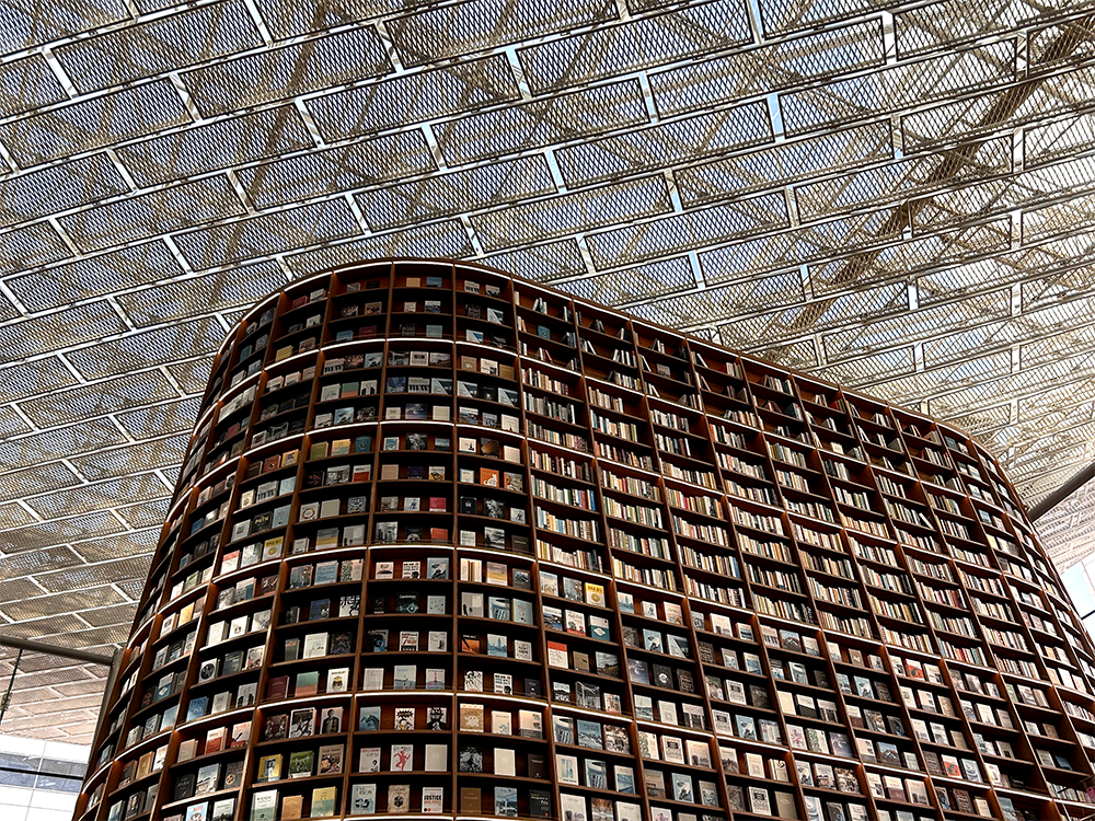 Photograph of a large, curved bookcase, reaching up toward a ceiling made of a metal grid, through which the sky appears