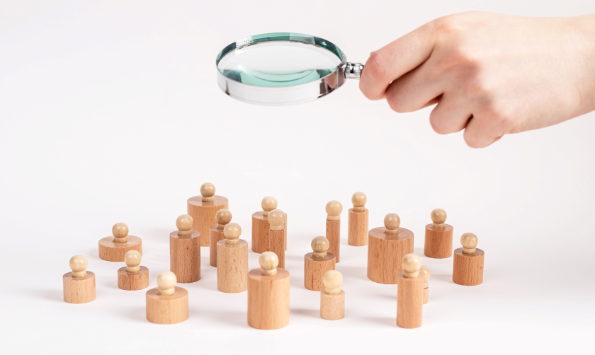 A photo illustration of a hand holding a magnifying glass above a group of wooden-peg people 