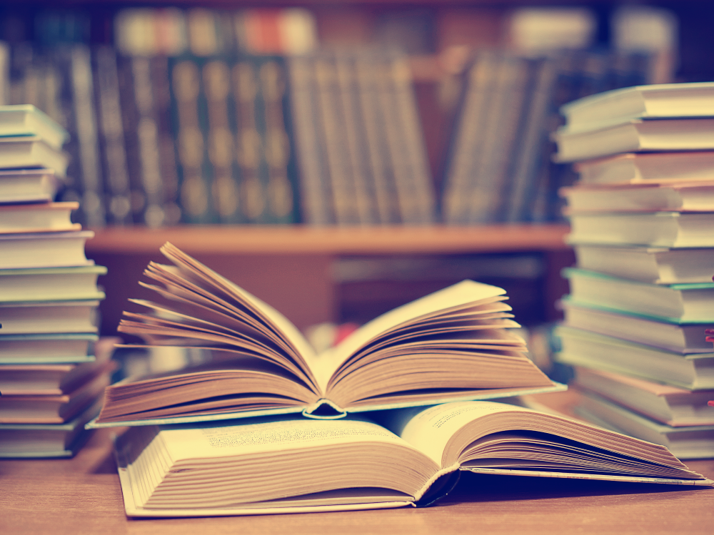 A small stack of open books, flanked by larger stacks of books, with shelves books in the background.