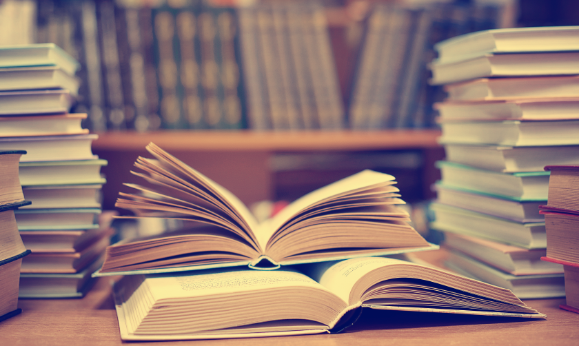 A small stack of open books, flanked by larger stacks of books, with shelves books in the background.