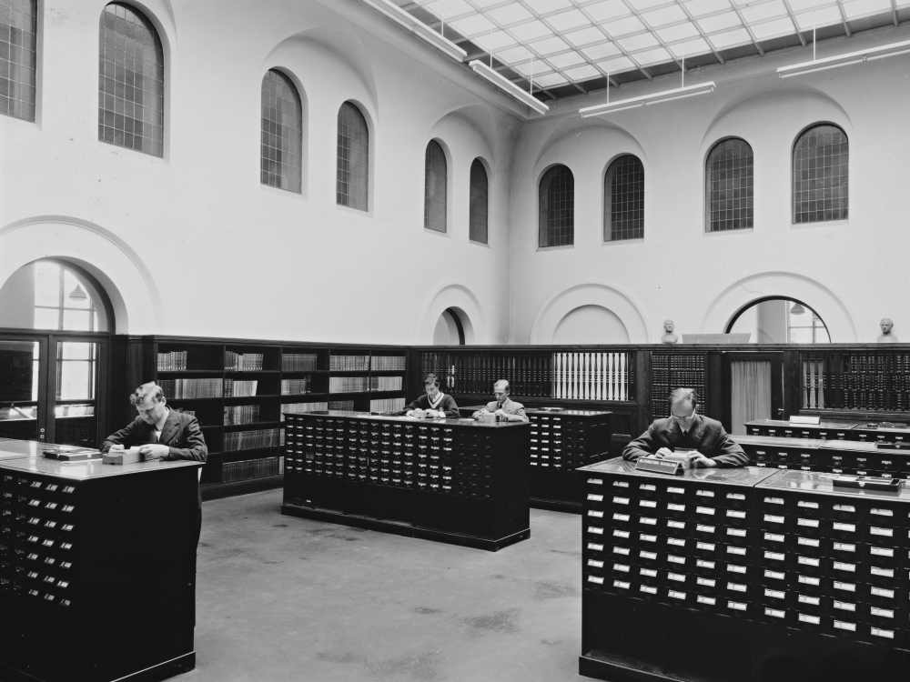 Black and white photograph of four men inside a library looking up information in a card catalogue.