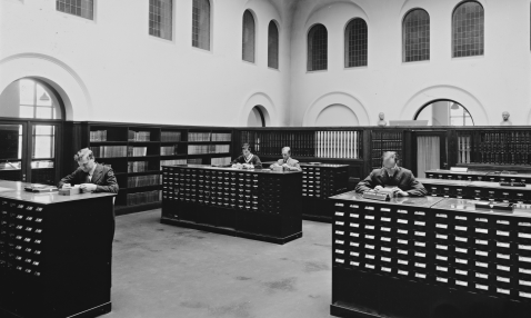 Black and white photograph of four men inside a library looking up information in a card catalogue.