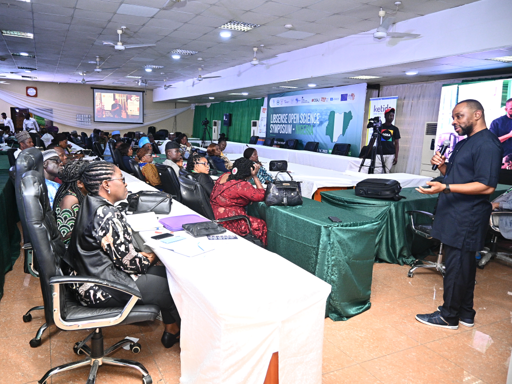 A photograph of a man, standing, addressing an audience seated behind long tables, with a banner at the front of the room reading “LIBSENSE OPEN SCIENCE SYMPOSIUM”