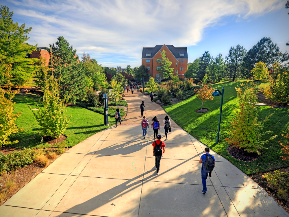 Students walk toward brick buildings along a sidewalk lined by green lawns and trees turning autumn colors 