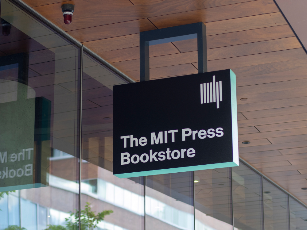 A black sign reading “The MIT Press Bookstore” in white text against a black background hanging in front of a wall of windows 