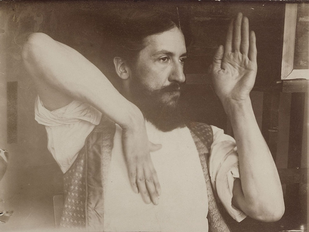 A scan of an old, slightly damaged photograph showing a bearded man with one elbow up in the air, hand down by his chest, and the other hand raised in front of his face.