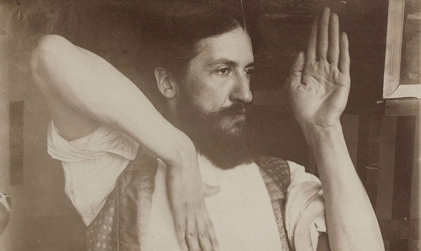 A scan of an old, slightly damaged photograph showing a bearded man with one elbow up in the air, hand down by his chest, and the other hand raised in front of his face.