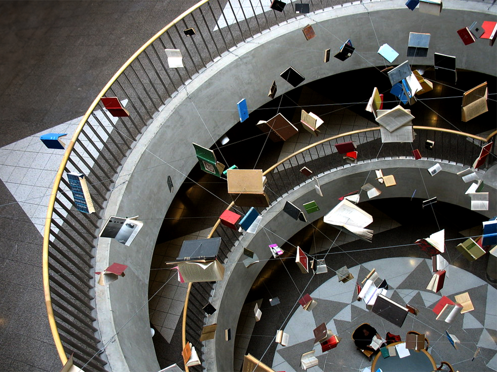 Photo of an art installation at the center of a circular stairwell featuring books suspended, open, on wires, with people reading in chairs at the edges