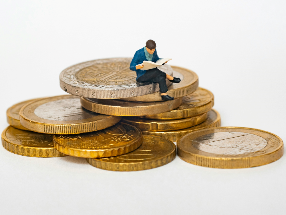 A man sits atop a pile of enormous coins, reading from a newspaper or magazine.