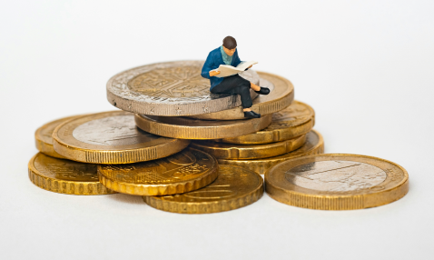 A man sits atop a pile of enormous coins, reading from a newspaper or magazine.