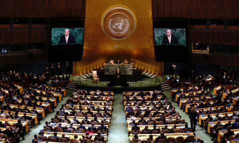 Photograph of a large room with full rows of seats facing a podium where a man is speaking; behind him are people seated at a desk and his image projected on screens 
