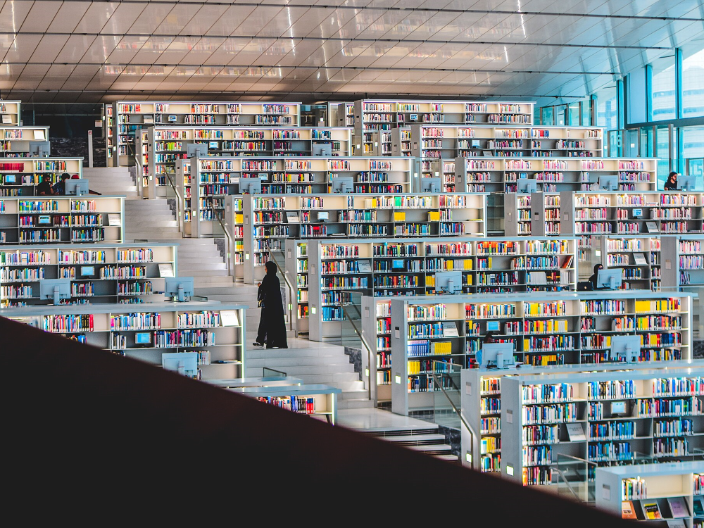 Photograph of rows of bright, white library shelves, surrounding a central staircase, on which a woman stands, browsing. 