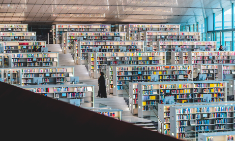 Photograph of rows of bright, white library shelves, surrounding a central staircase, on which a woman stands, browsing. 