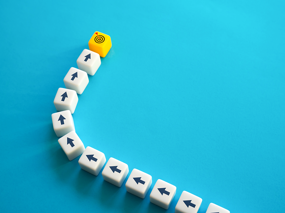 Photo of a series of cubes with arrows pointing forward on them arranged in a curved line ending at a different colored cube with an image of an arrow hitting a target on it
