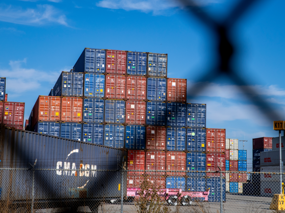A photograph of a stack of shipping containers viewed through a chainlink fence 