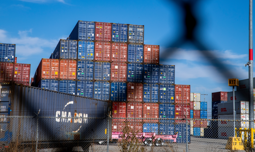 A photograph of a stack of shipping containers viewed through a chainlink fence 