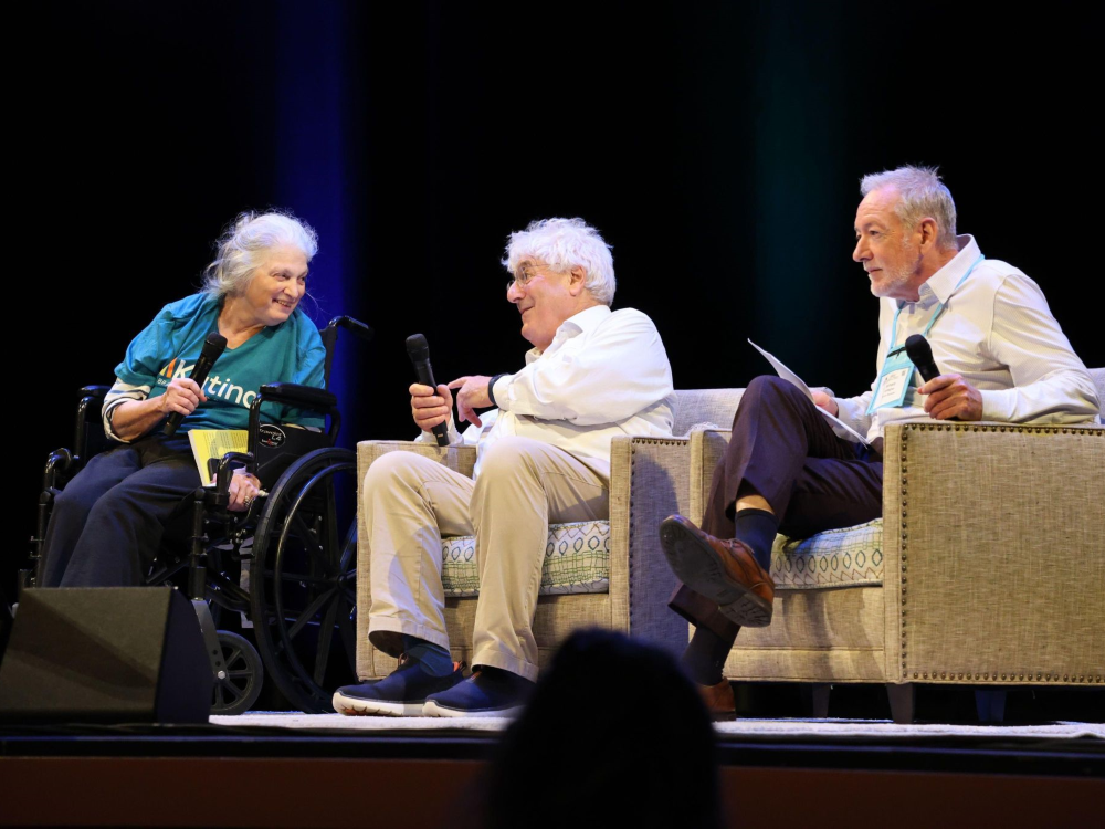 Three people seated in a row, on a stage, with microphones, engaged in conversation