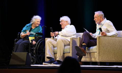 Three people seated in a row, on a stage, with microphones, engaged in conversation