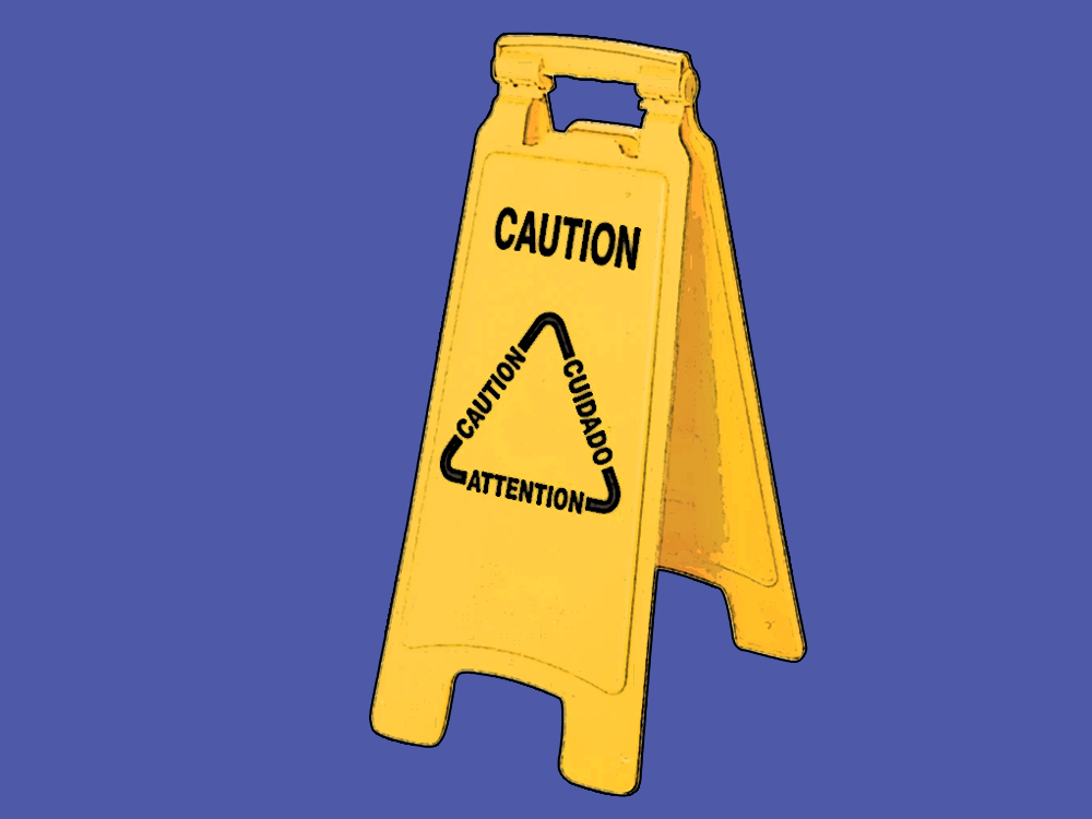 A yellow stand up floor sign reading “CAUTION” at the top and “Caution” “Cuidado” and “Attention” on the three sides of a triangle at the center 