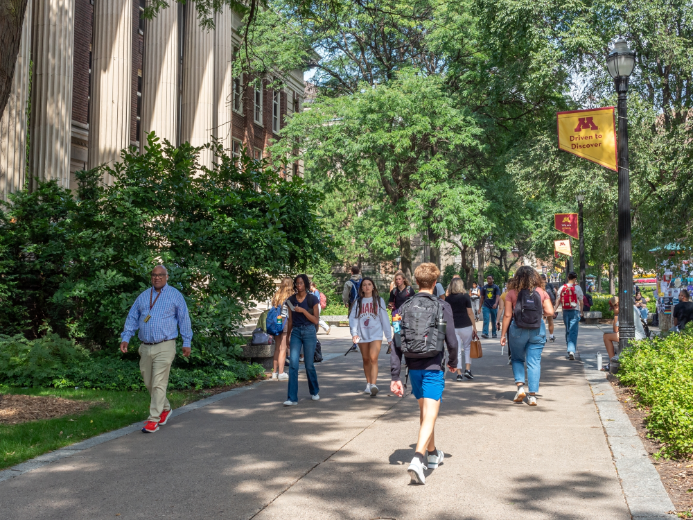 A photo of people walking on a wide sidewalk past a university building and lightpoles with university banners mounted on them