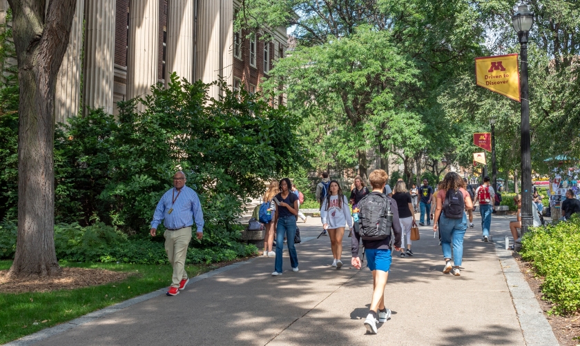 A photo of people walking on a wide sidewalk past a university building and lightpoles with university banners mounted on them