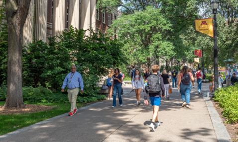 A photo of people walking on a wide sidewalk past a university building and lightpoles with university banners mounted on them