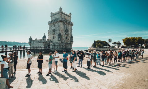 A line of tourists in front of an old looking stone tower in front of the sea