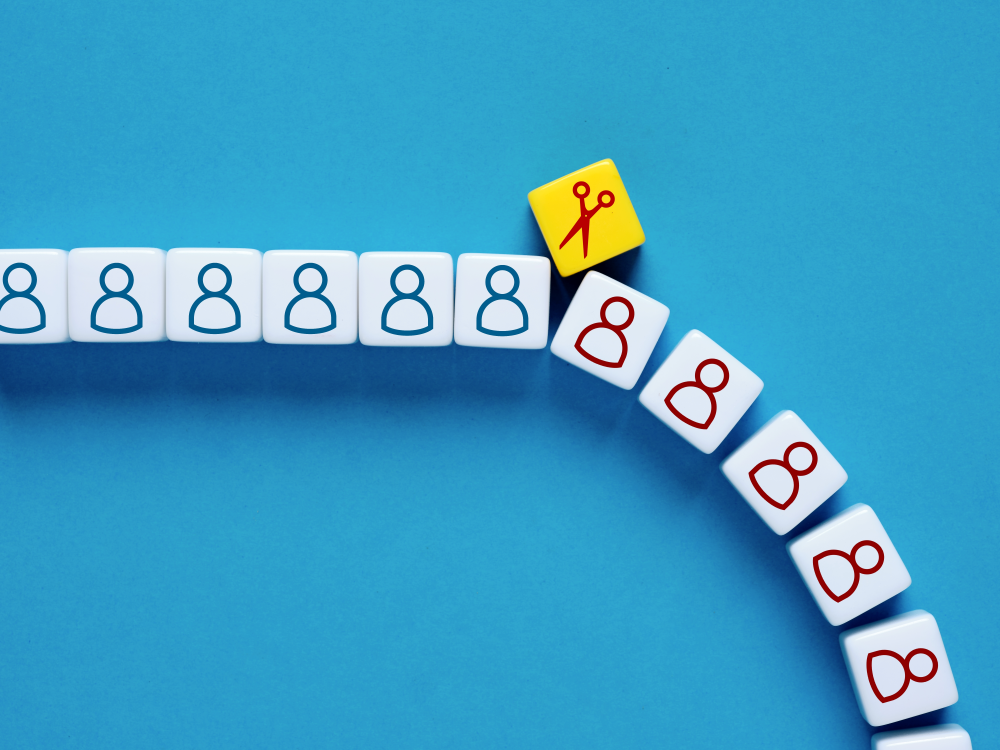 A row of cubes with outlines of people on them, divided by a cube with a pair of scissors on it, after which the people are red instead of blue and the row slants downward, as if they cubes are falling. 